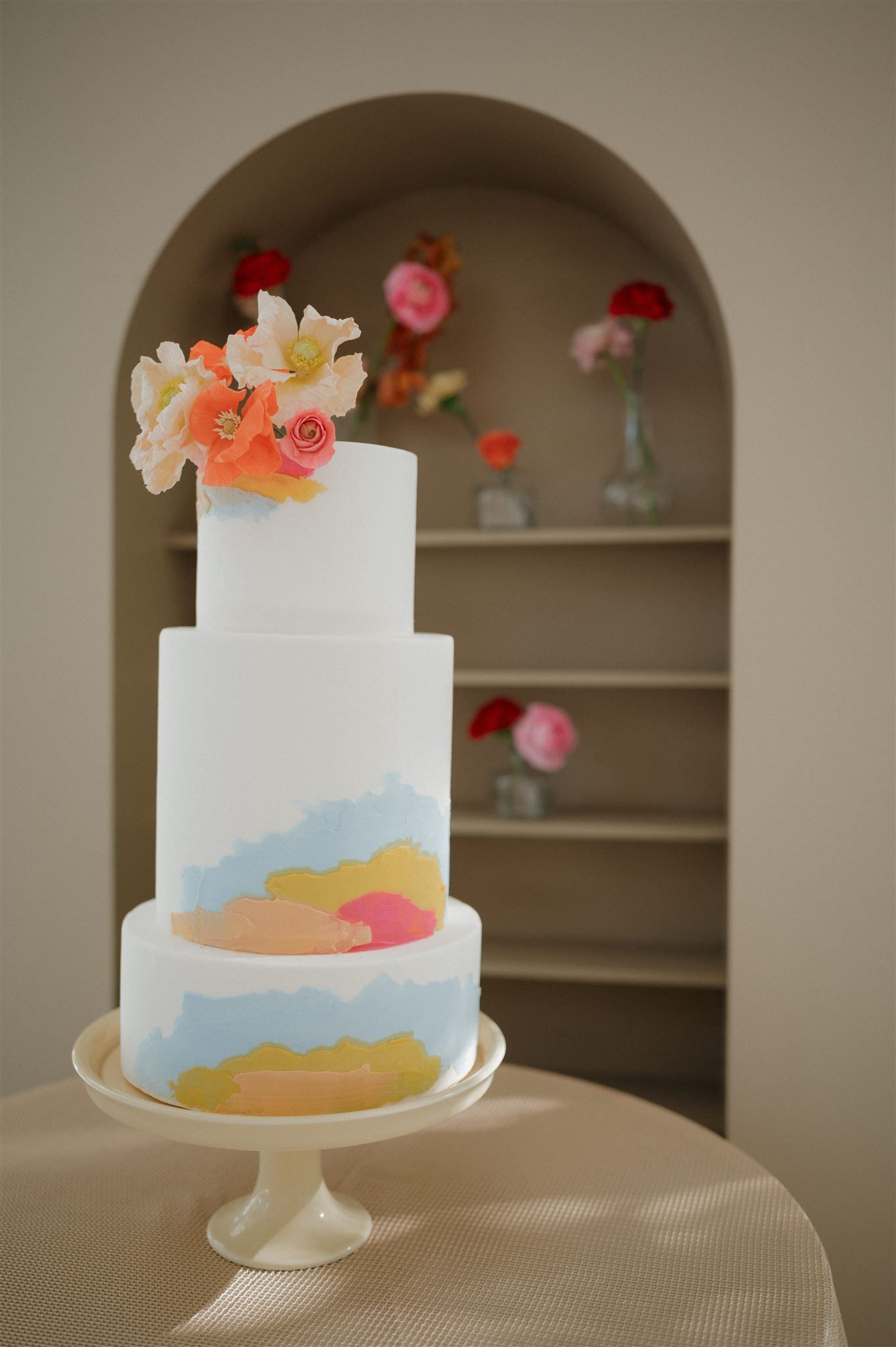 Three-tiered white wedding cake with watercolor-style pastel design and fresh floral decorations displayed in an arched nook.