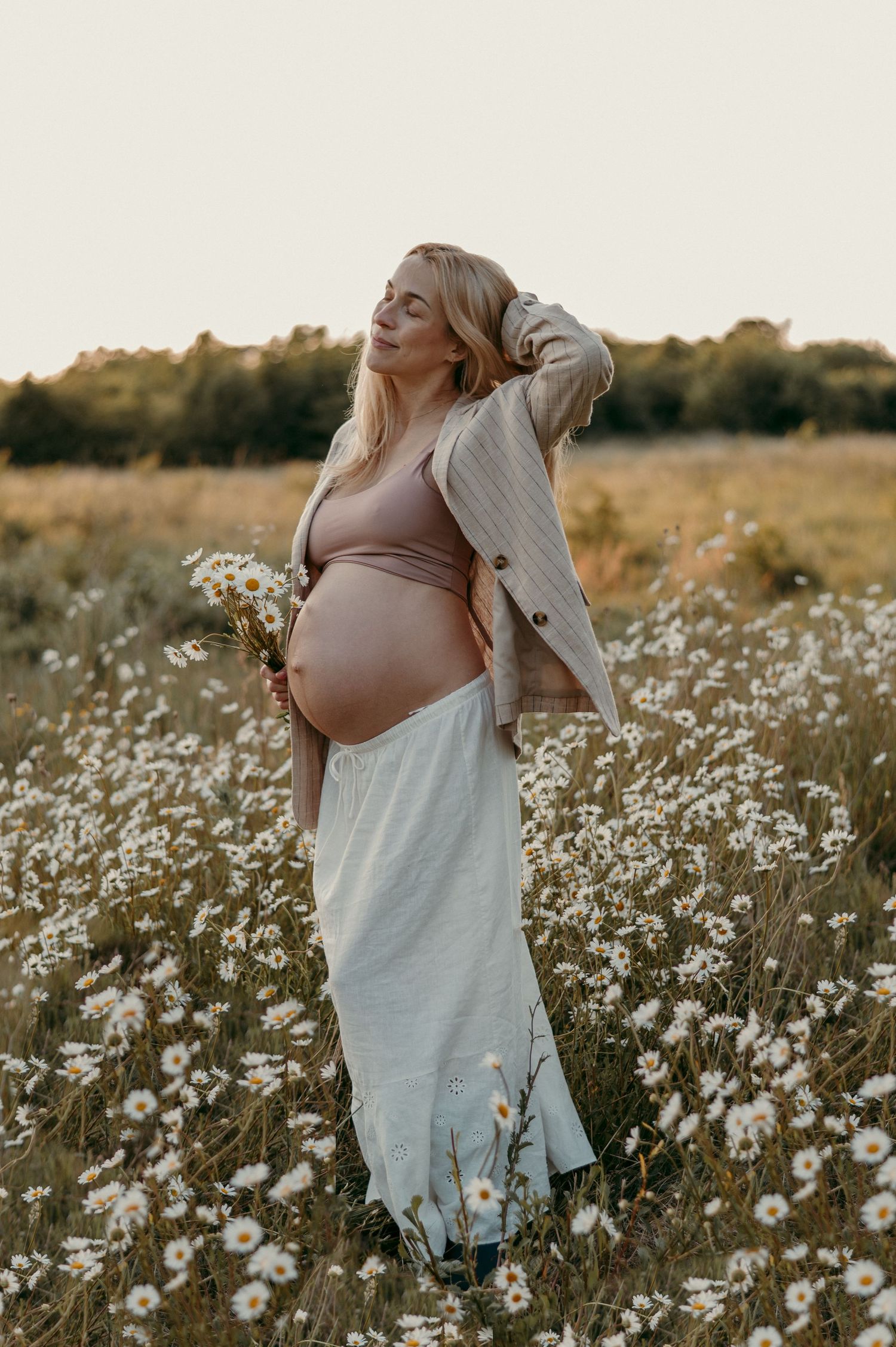 Maternity photoshoot in a field of white wildflowers during golden hour sunset.