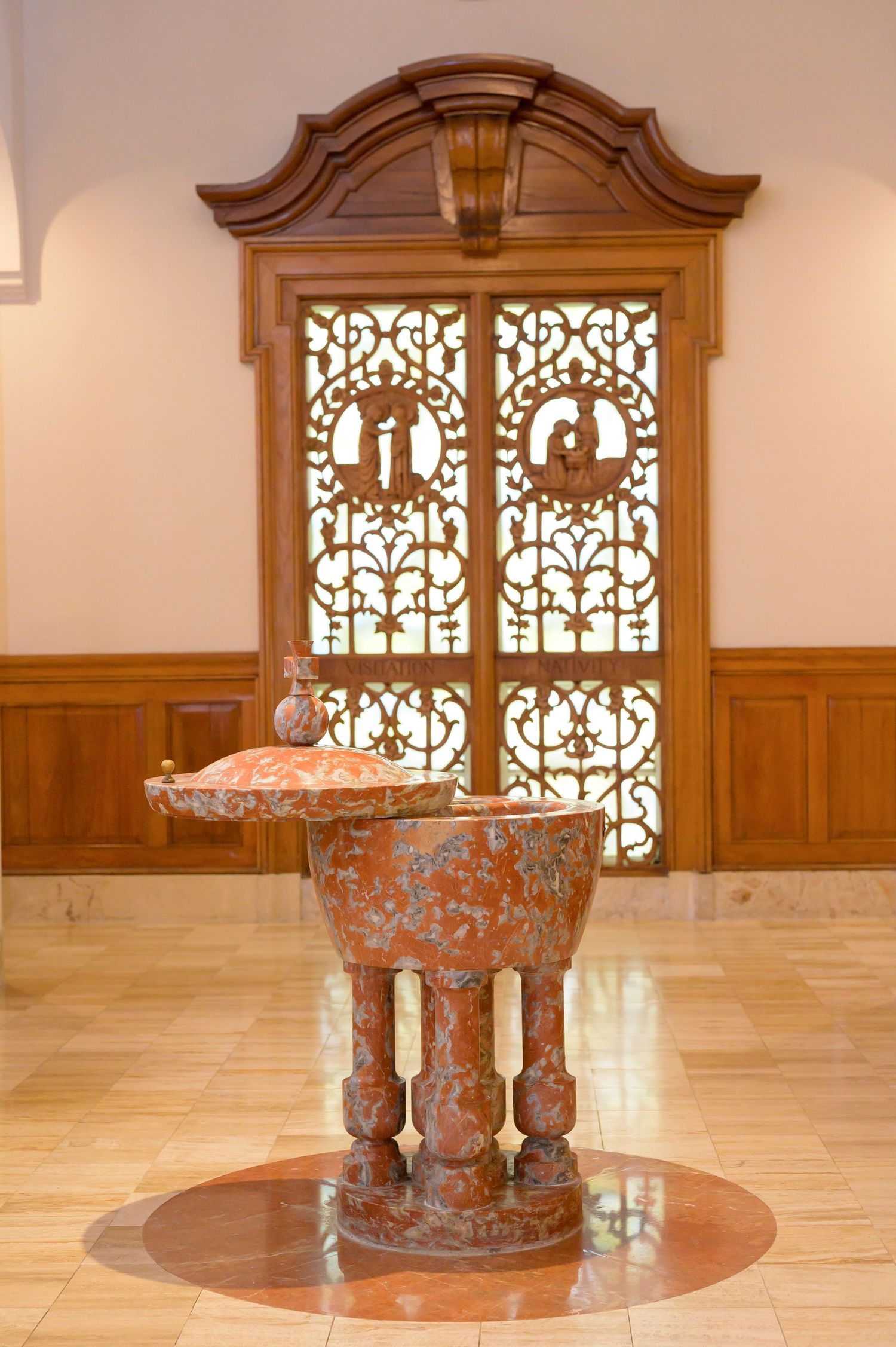 Ornate baptismal font made of marble stands in front of decorative wooden doors with intricate lattice patterns.