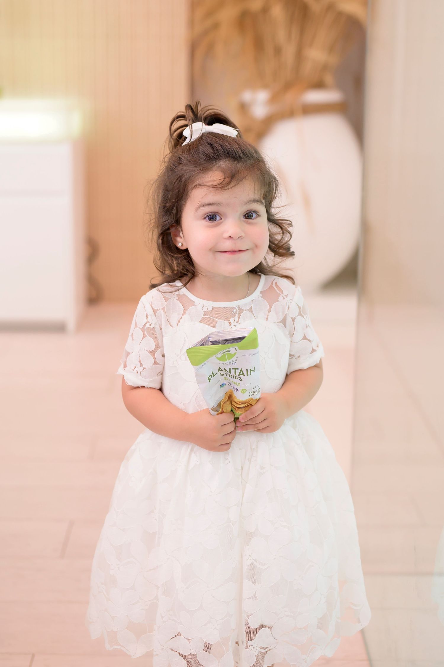 Sweet toddler in white lace dress holding a snack package stands in room with soft lighting and decor.