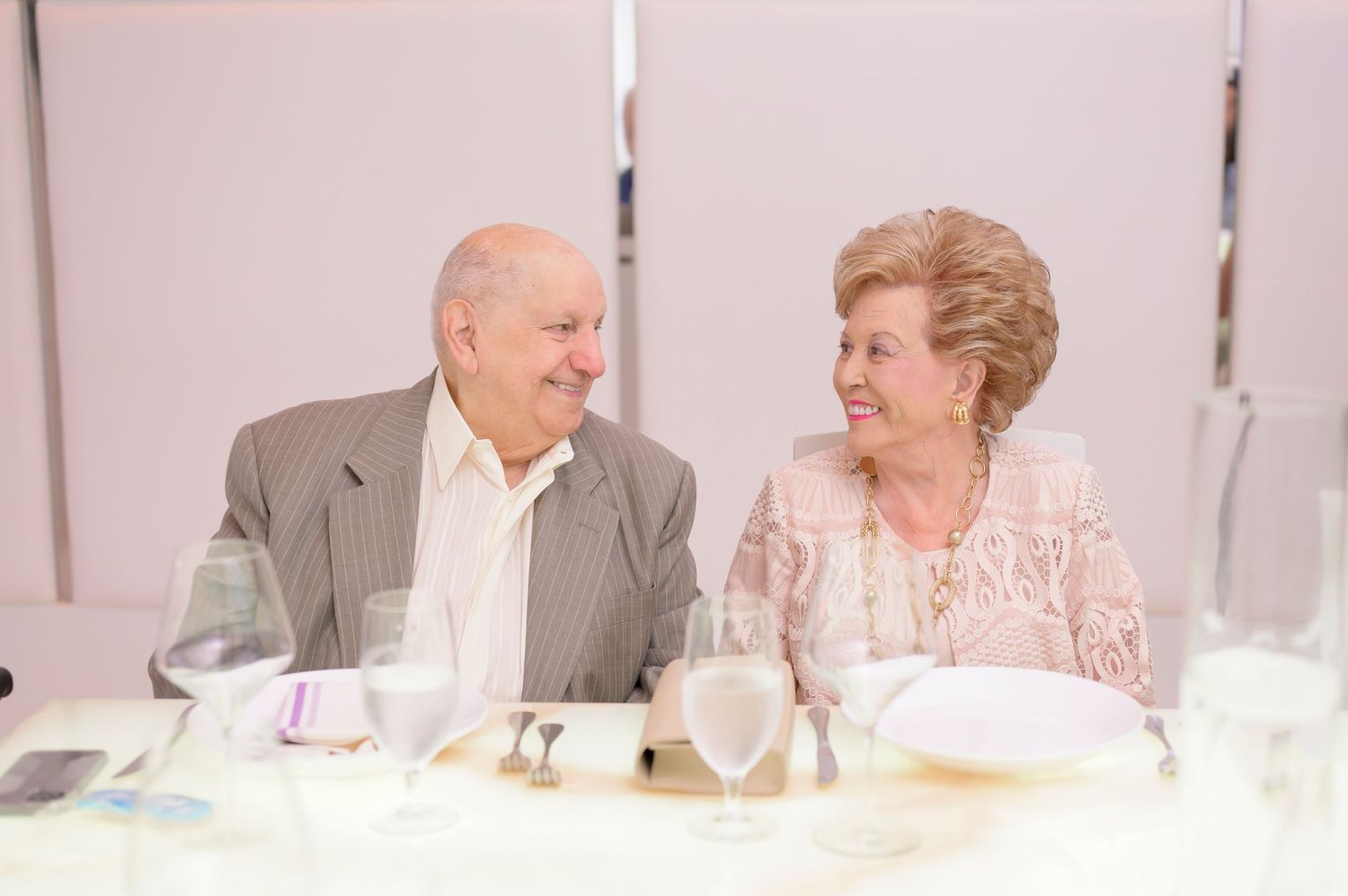 An elderly couple shares a joyful moment while sitting together at a formal dining table.