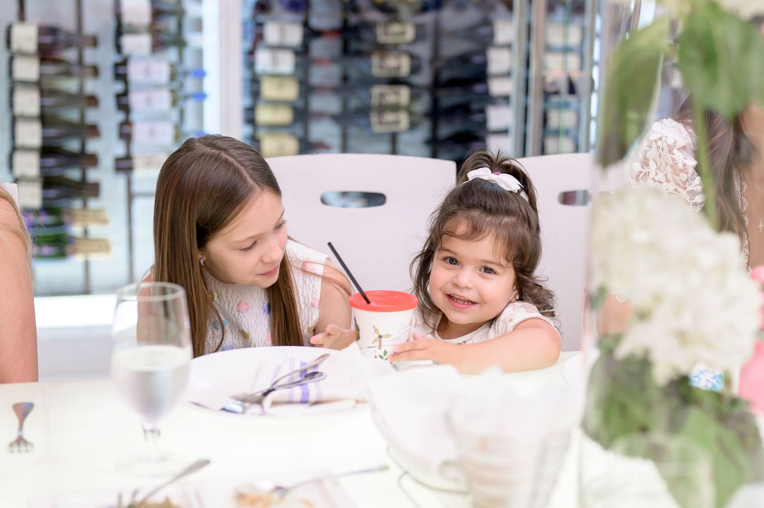 Two young children sitting at a restaurant table with white tablecloths and glassware during a special occasion.