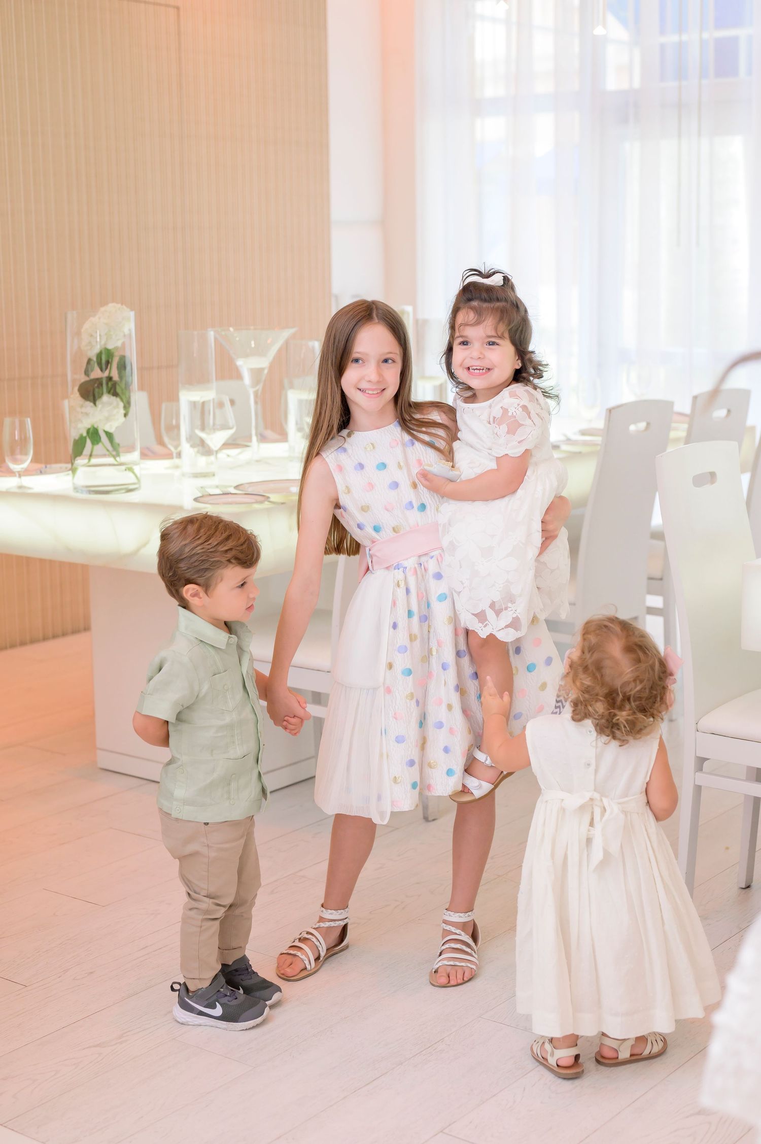 A sunny indoor gathering shows a group in coordinated light outfits standing together in an elegant white dining room setting.