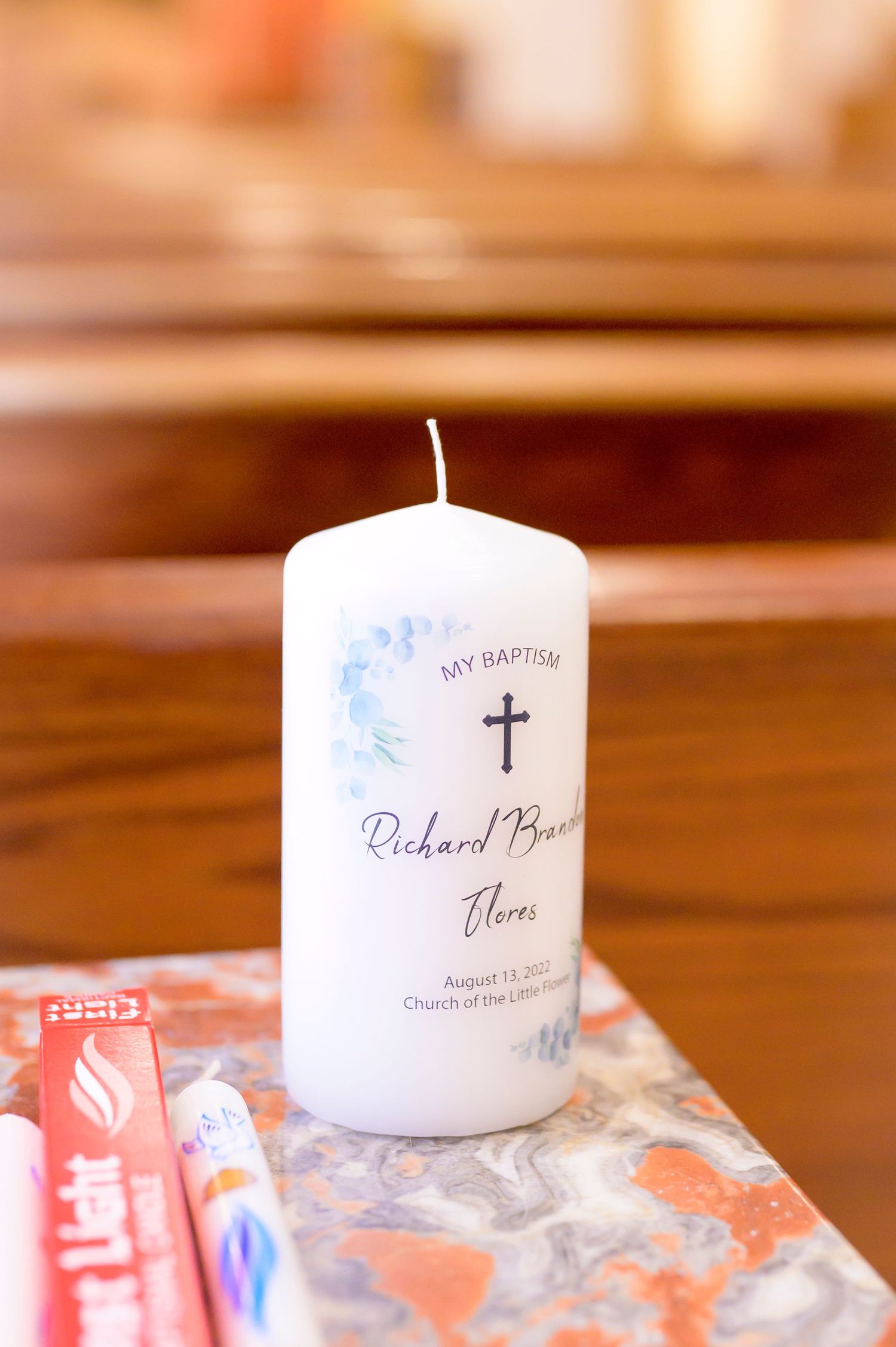A white baptismal candle with a cross symbol and handwritten text sits on a wooden church pew.