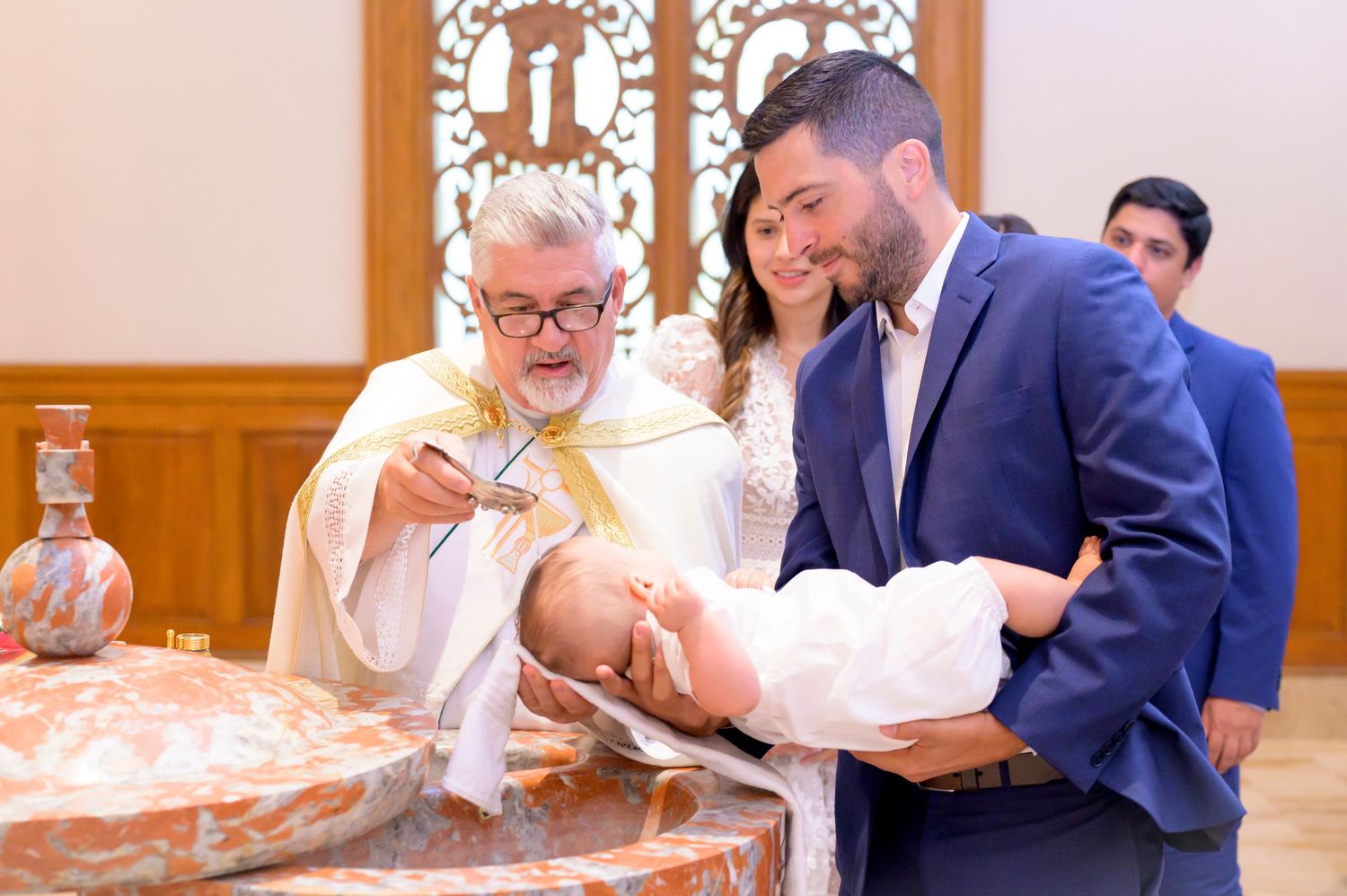 A priest performs a baptism ceremony while wearing traditional vestments in a church with wooden paneling.