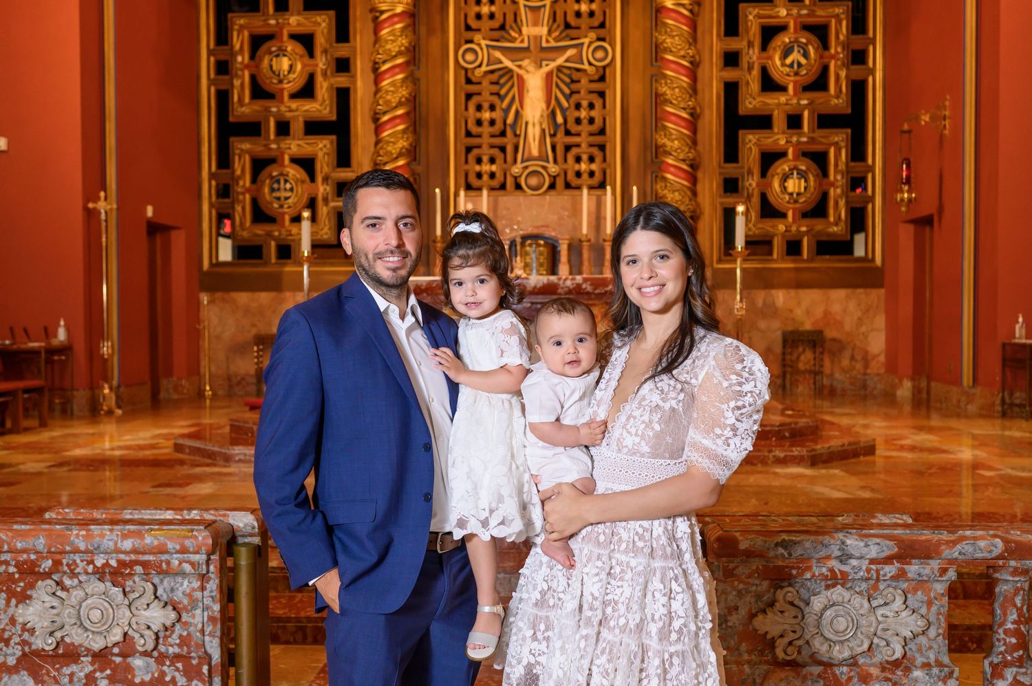 A family in formal attire poses together against an ornate wooden backdrop with decorative patterns and rich red tones.