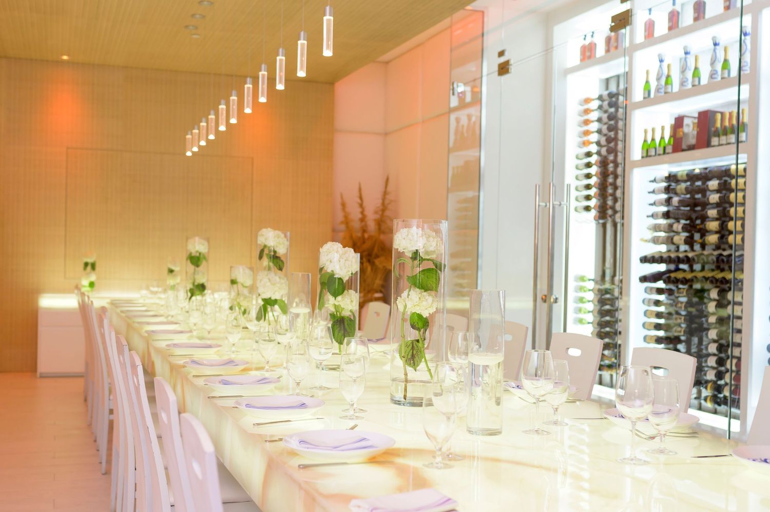 An elegant white dining table set for a formal event with floral centerpieces and wine displays along the wall.
