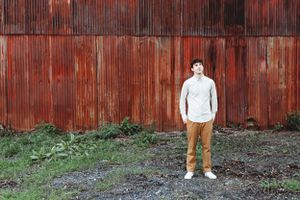 Person in white shirt and tan pants standing against rustic red wooden wall.