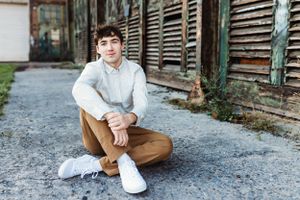 Casual portrait of graduating senior man in white shirt and brown pants sitting on concrete ground.