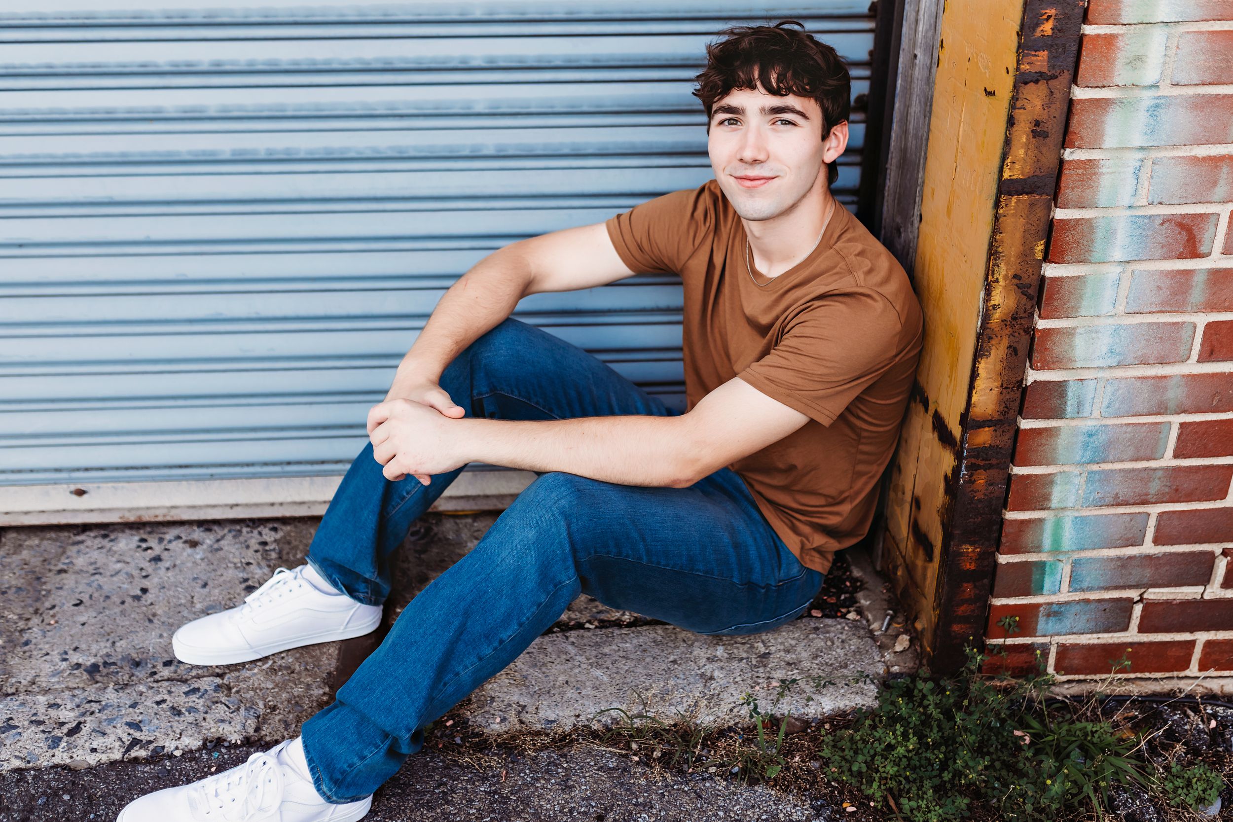 Person in brown shirt and jeans sitting casually against industrial doorway.