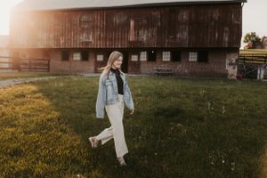 Person in denim and white outfit walking in front of vintage barn at sunset.
