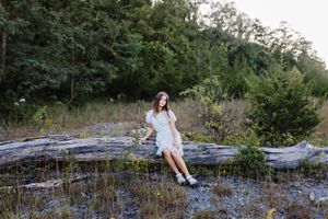 Someone in a white dress sitting on a fallen log surrounded by wildflowers and trees.