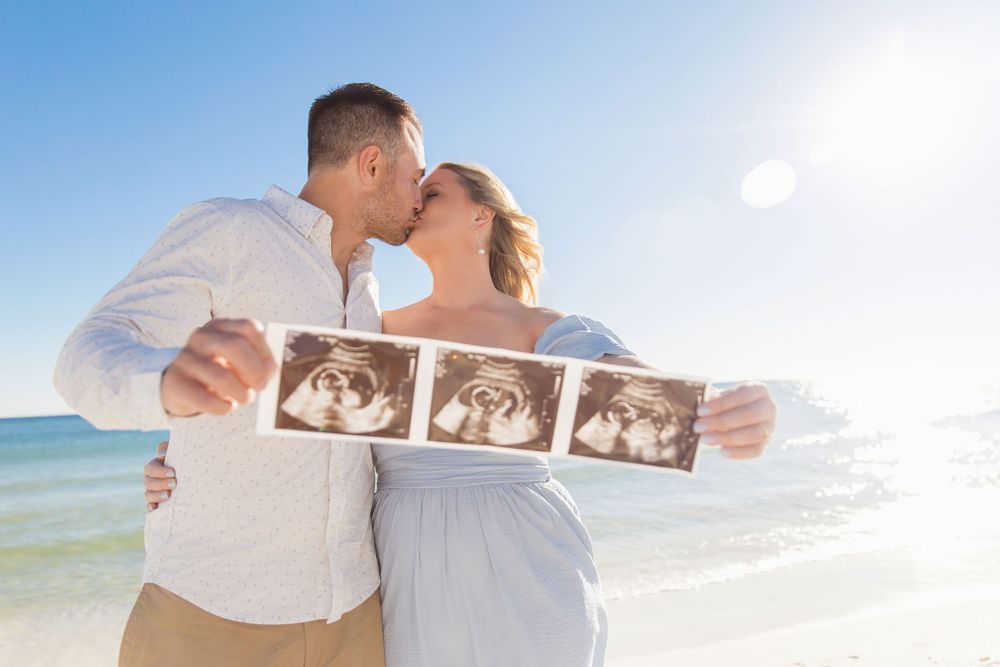 Couple sharing intimate moment on beach while displaying ultrasound photos.