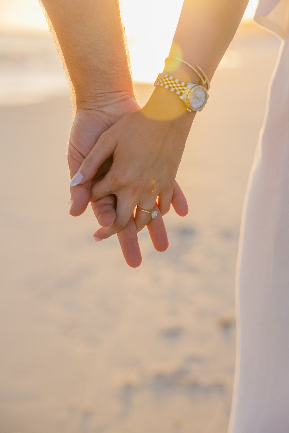 Close-up view of hands holding each other against a sandy beach background at sunset.