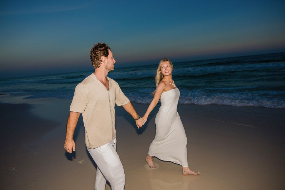 Couple in white and beige outfits walking along sandy beach during golden hour.