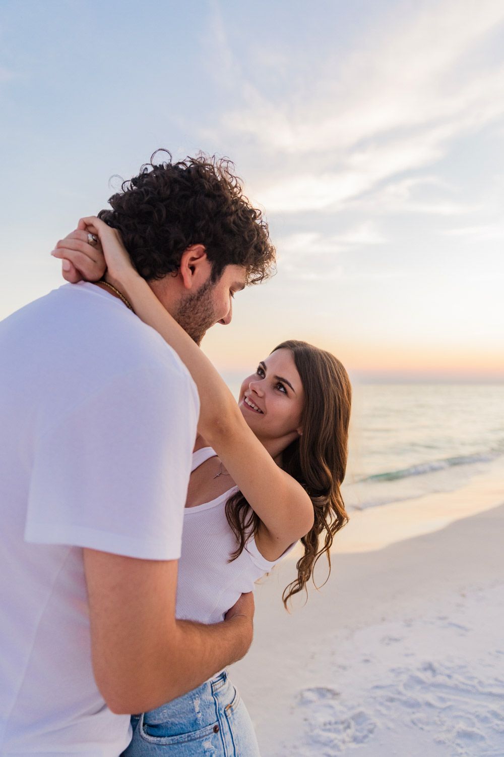 A couple dances together on a white sandy beach at sunset with soft pastel clouds in the background.