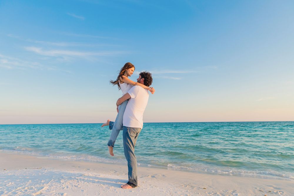 Woman in white shirt and jeans walking along pristine white sand beach with turquoise ocean waters on sunny day.