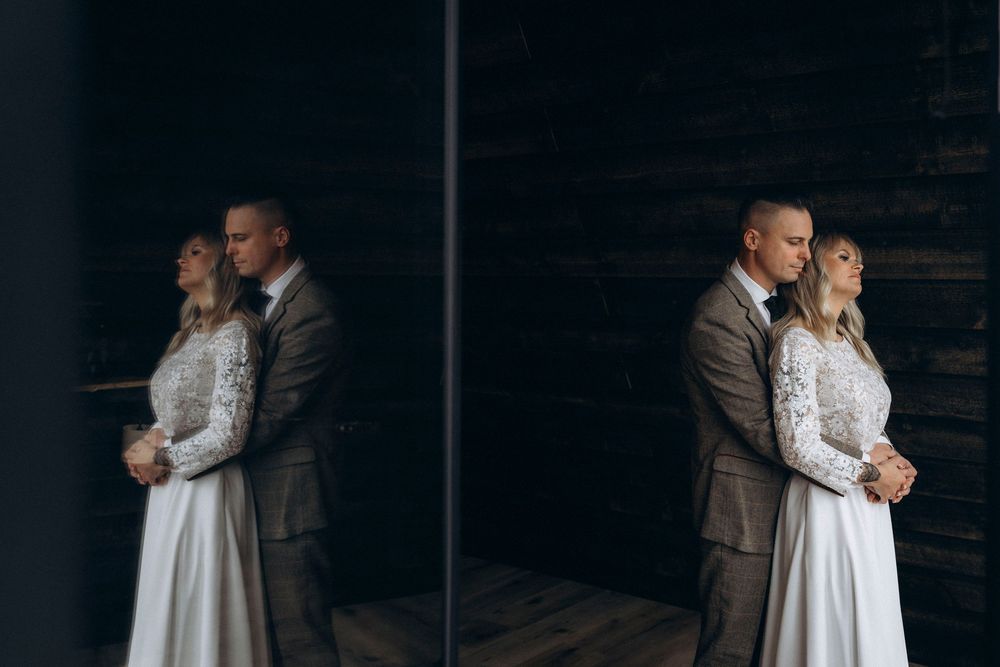 Artistic wedding portrait showing couple embracing in white dress and gray suit with mirrored reflection against dark wall.