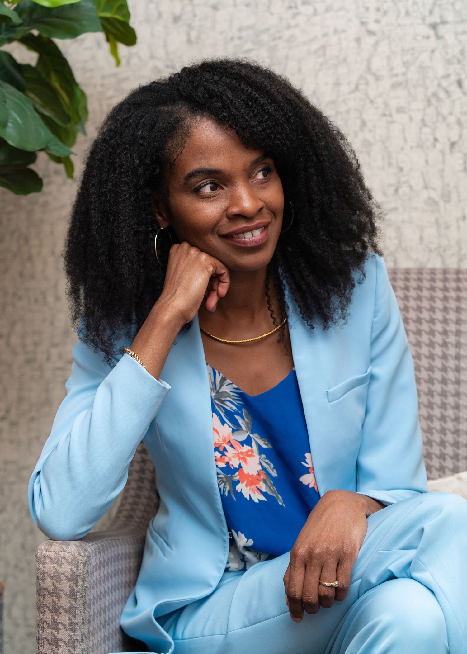 Stylish Black woman in light blue blazer smiling while seated in a bright outdoor setting.