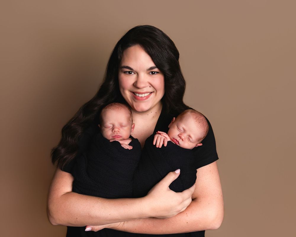 Smiling mother in black dress holding two sleeping newborn twins wrapped in black swaddles against a brown background.