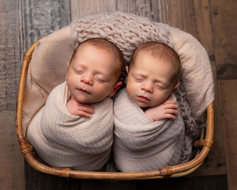Two swaddled newborn twins sleeping peacefully side by side in a wicker basket with soft blankets.