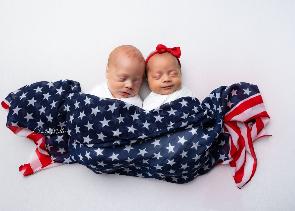 Two sleeping infants wrapped together in a patriotic American flag themed blanket with stars and stripes pattern.