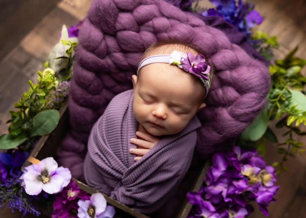 Newborn baby wrapped in purple swaddle sleeping in a wooden bowl surrounded by purple and green flowers.