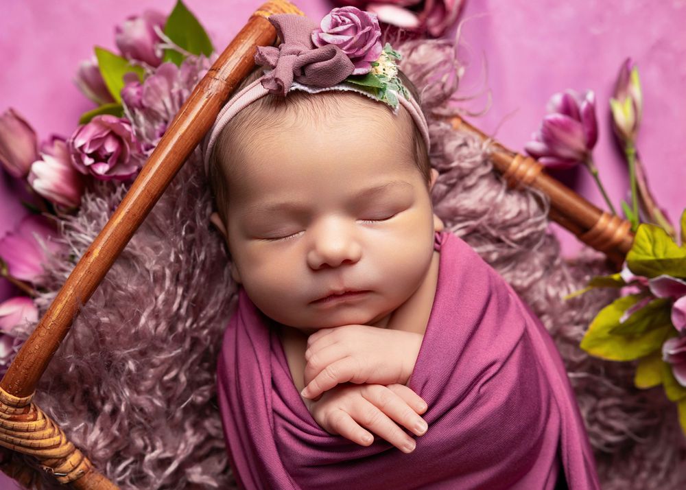 Newborn baby swaddled in purple wrap sleeping in a basket with floral headband and pink flowers.