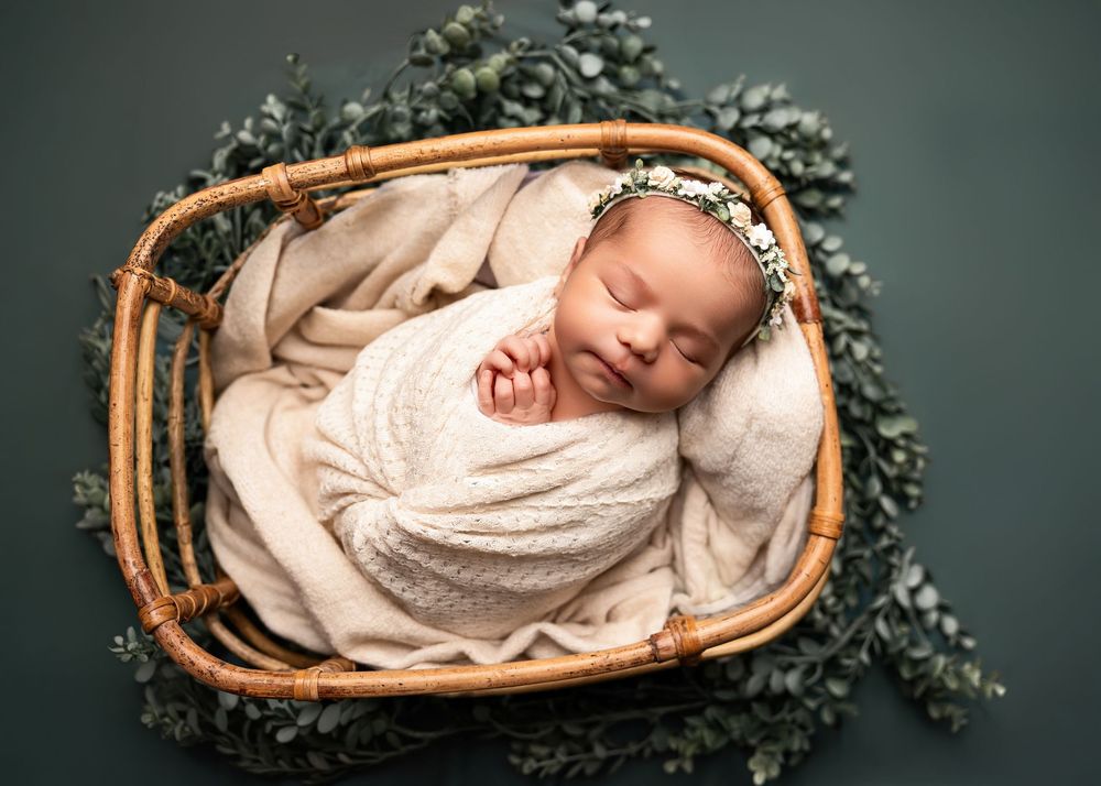 Sleeping newborn baby wrapped in cream knit blanket in wicker basket with eucalyptus, wearing floral headband.