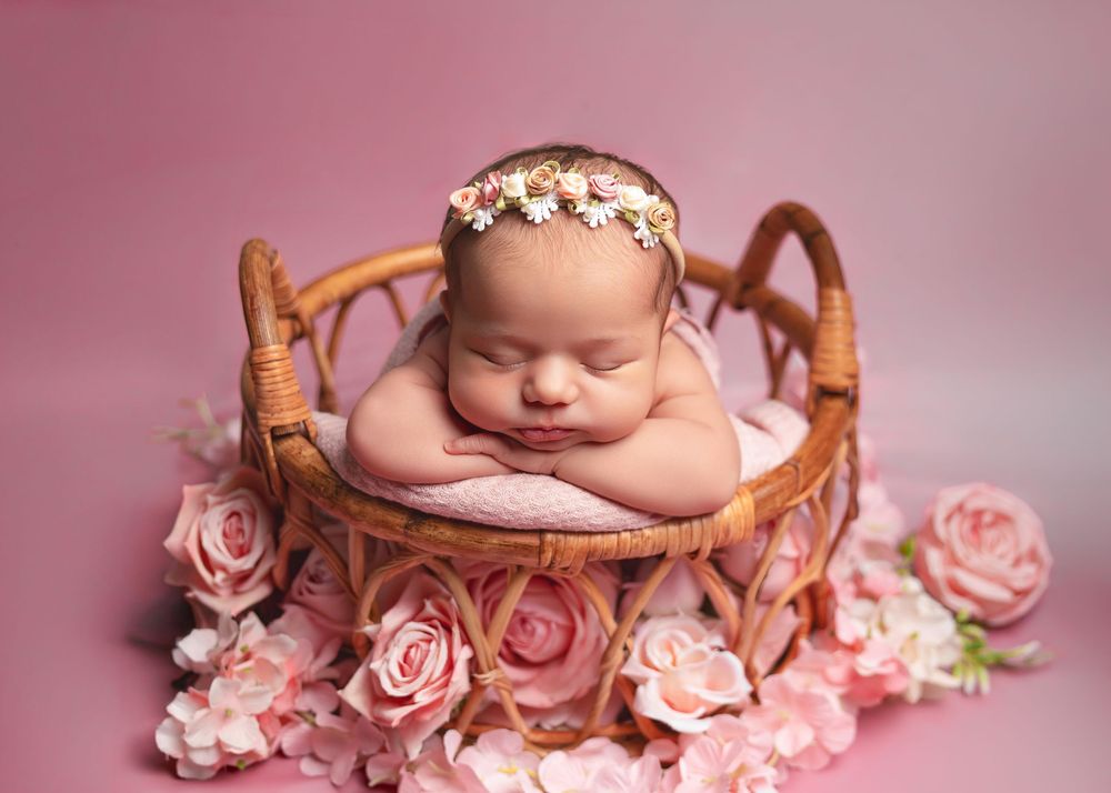 Sleeping newborn baby in floral headband posed in wicker basket surrounded by pink roses on pink background.