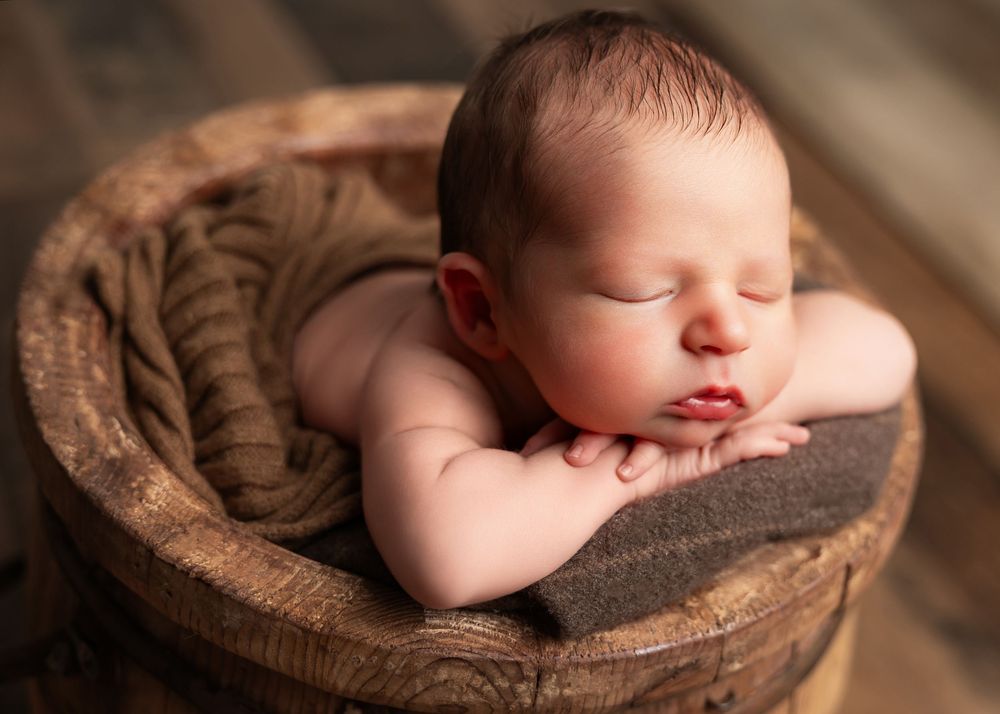 Newborn baby sleeping peacefully in a rustic wooden bucket with a soft brown knit wrap.