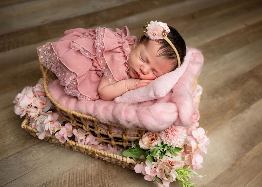 Newborn baby girl sleeping in wicker basket with pink blanket, floral headband, and pink dress surrounded by flowers.