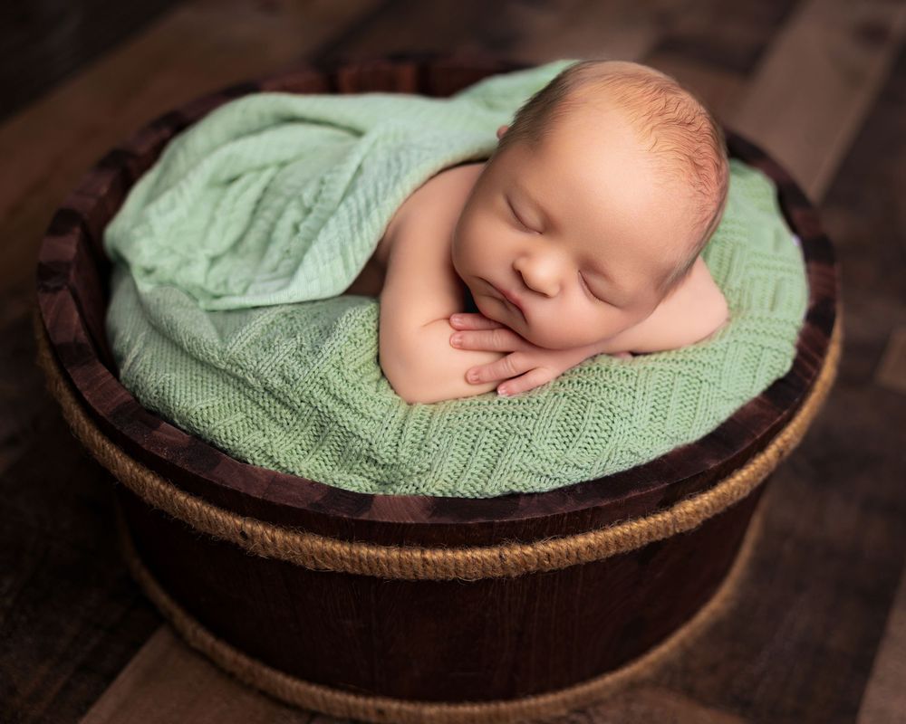 Sleeping newborn baby wrapped in a soft green knit blanket nestled inside a rustic wooden bowl prop.