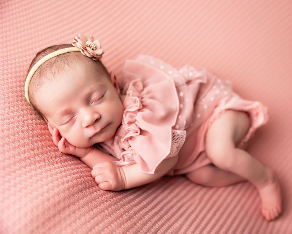 Newborn baby girl sleeping peacefully in pink ruffled outfit and floral headband on soft pink knit blanket.