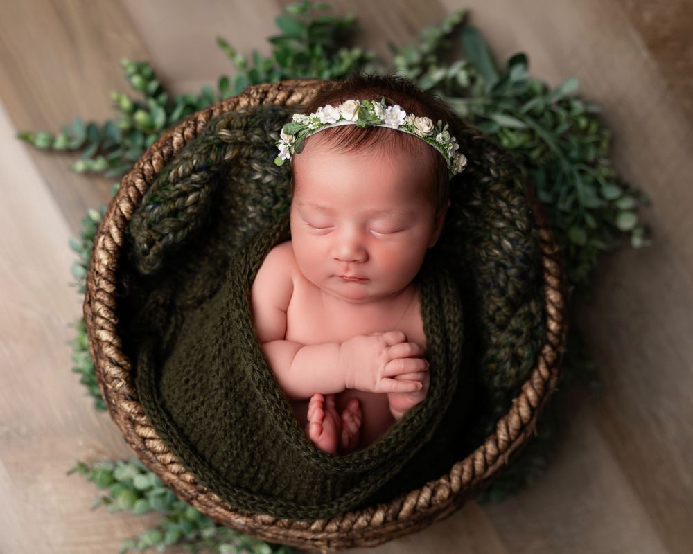 Newborn baby sleeping in a woven basket wrapped in green knit blanket with a floral crown on wood floor.