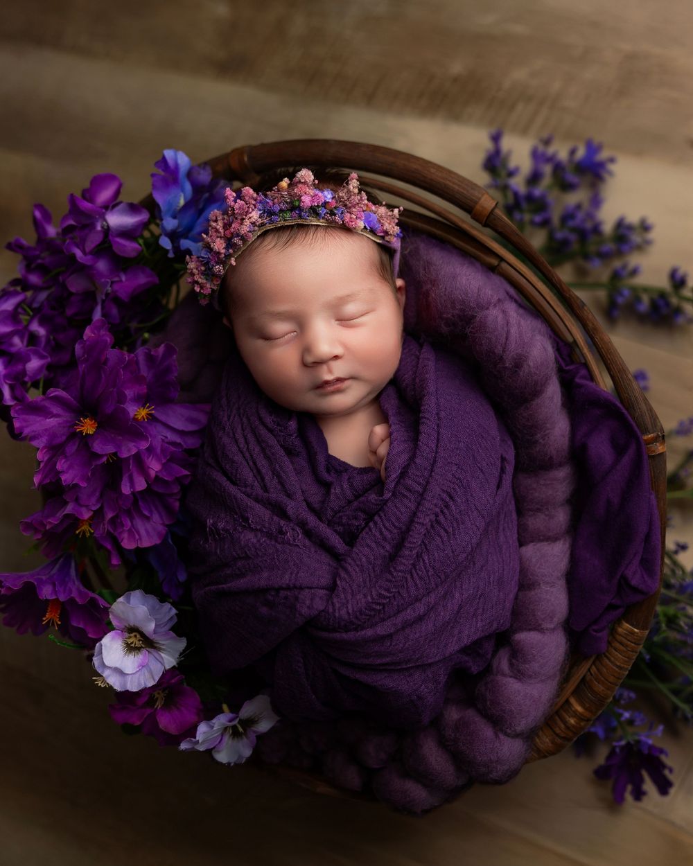 Newborn baby wrapped in purple swaddle with floral crown, nestled in wicker basket surrounded by purple flowers.