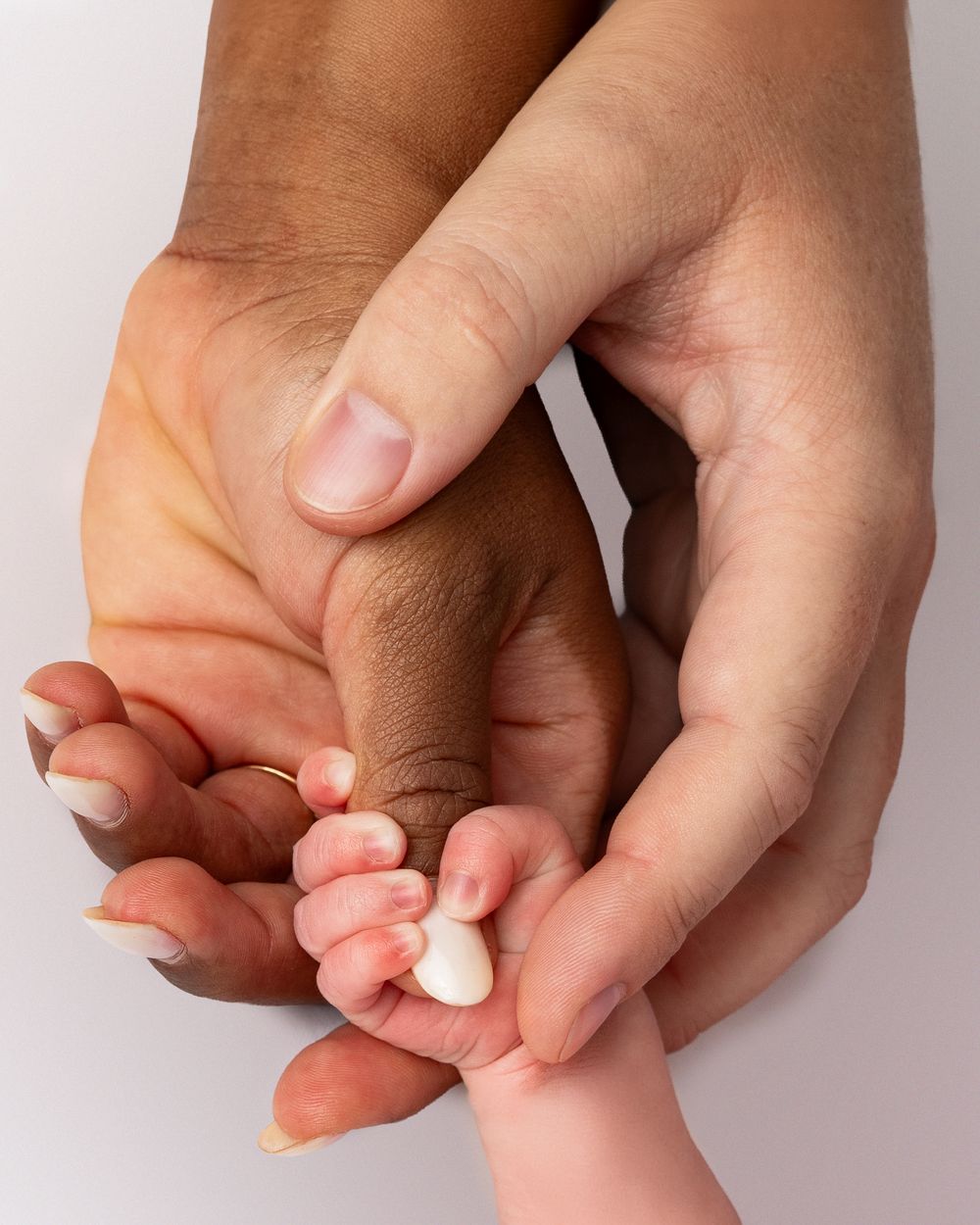 Two adult hands of different skin tones gently cradling a newborn baby's tiny hand in a tender moment.