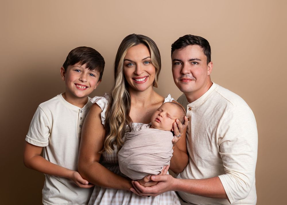 Family of four posing for newborn portrait, parents with young boy holding swaddled baby, neutral beige background.