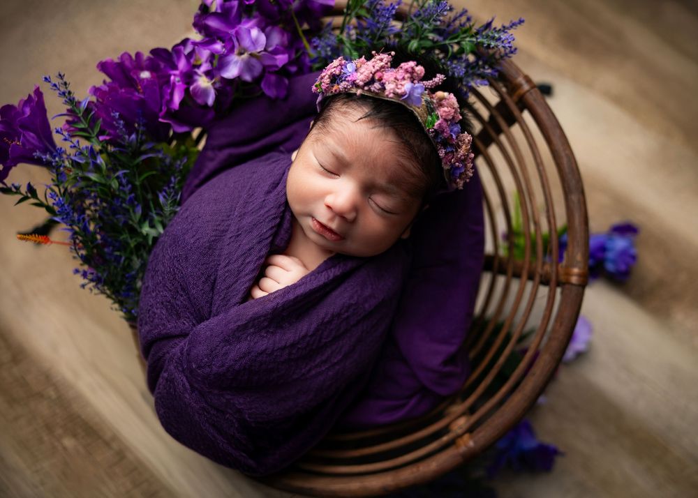 Newborn wrapped in purple blanket sleeping in wooden basket with purple and pink floral decorations.