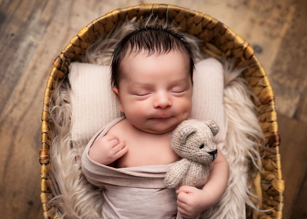 Smiling newborn baby wrapped in beige swaddle holds tiny knit teddy bear while sleeping in wicker basket with fur lining.