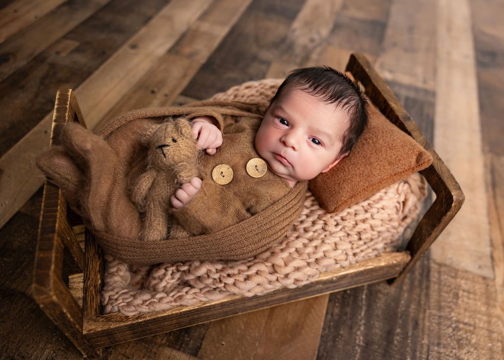 Newborn baby sleeping in a rustic wooden bed with brown knit wrap and teddy bear in a cozy studio photo.