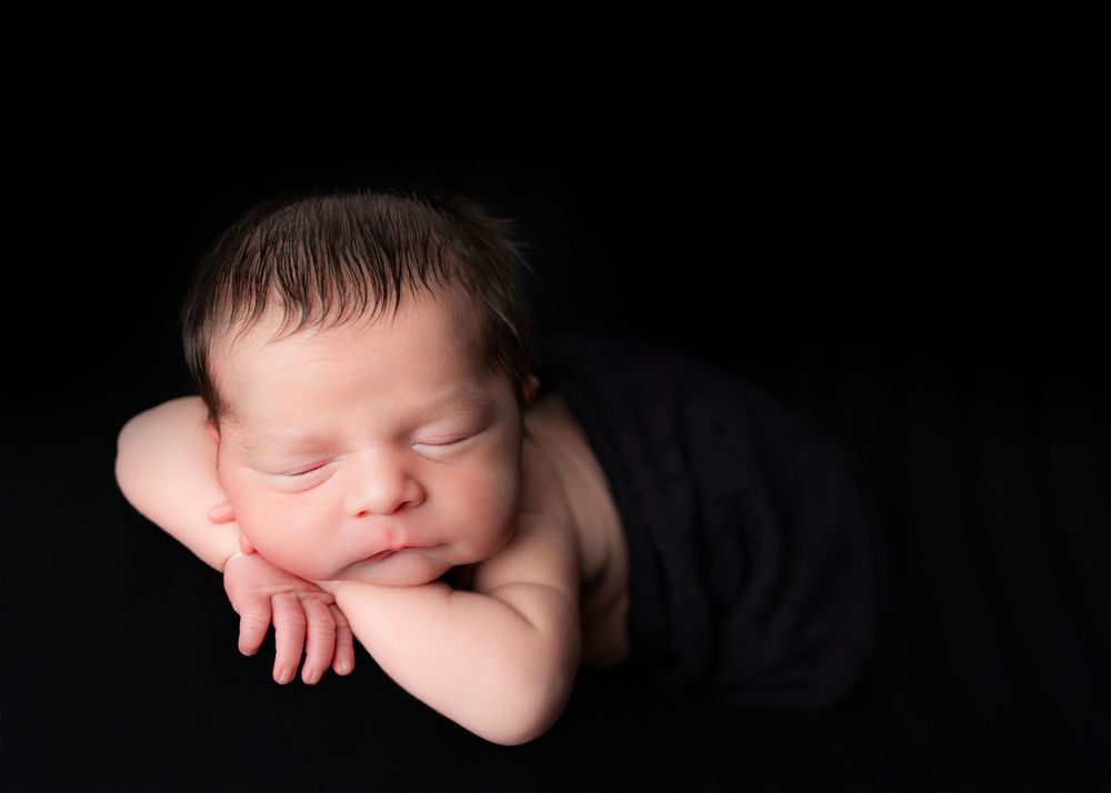Peaceful newborn baby sleeping on black background with hands tucked under chin in classic posed newborn photography.