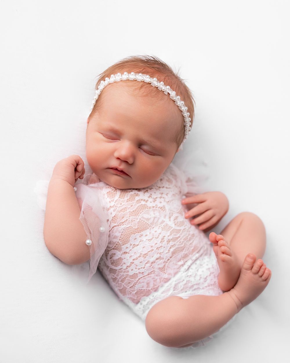 Sleeping newborn baby girl in white lace romper and pearl headband on white background.