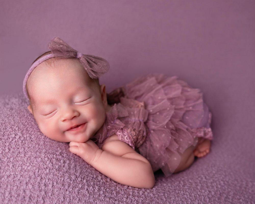 Smiling newborn baby in purple ruffled outfit and bow headband sleeping peacefully on lavender blanket.