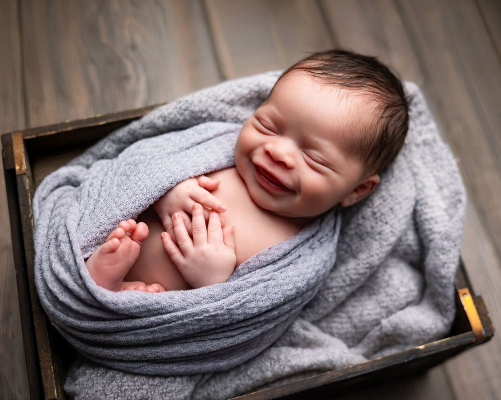 Smiling newborn baby wrapped in a soft gray blanket, nestled in a rustic wooden box on a wood floor.