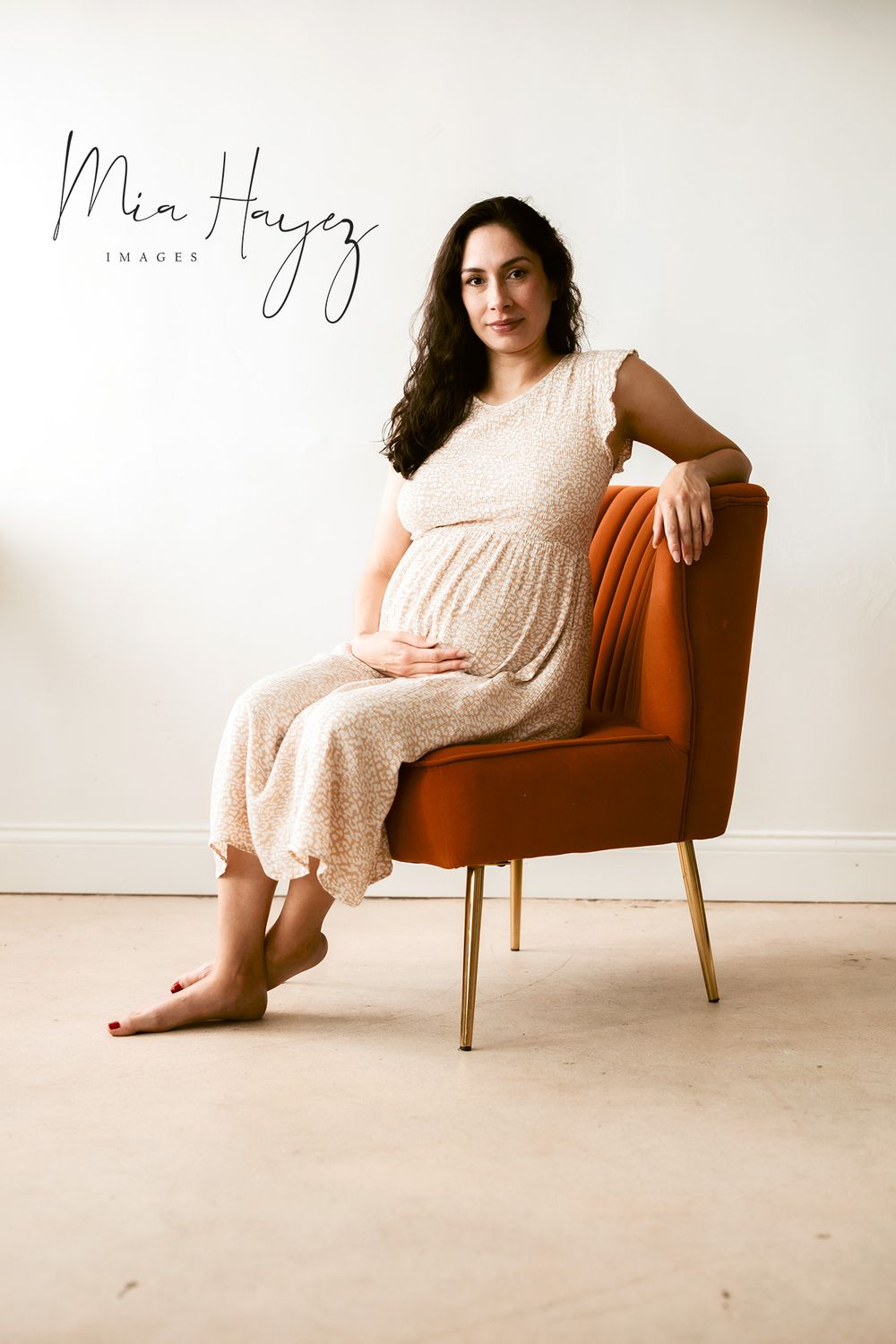 Artistic maternity portraits in a beige dress on a tan armchair against white walls.