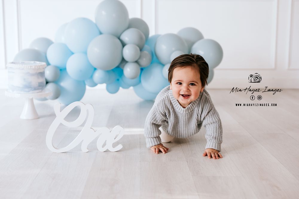 Baby milestone photoshoot with blue and grey balloon arch decoration on white backdrop.