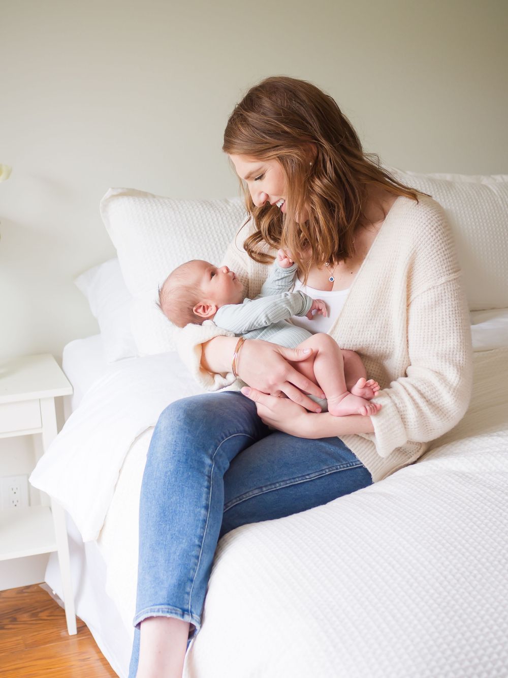 mom holding newborn baby in her arms while sitting on a white bed. plain white walls. light  and airy with sunshine. Sonoma County