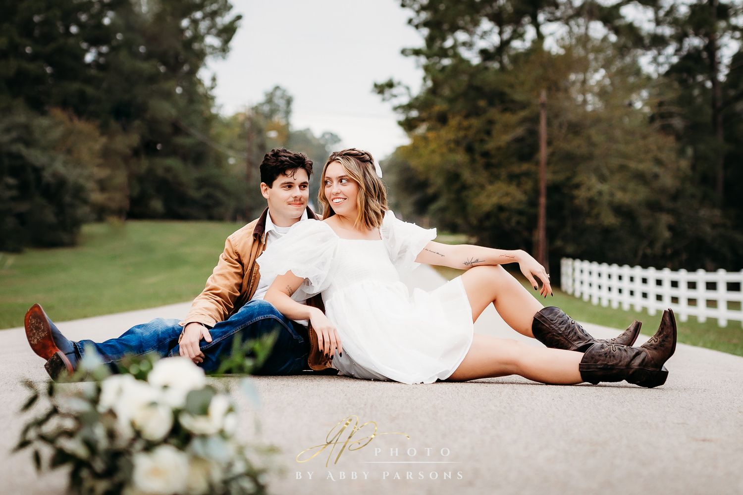 A romantic couple sitting together on a path near a white fence, surrounded by lush green trees.