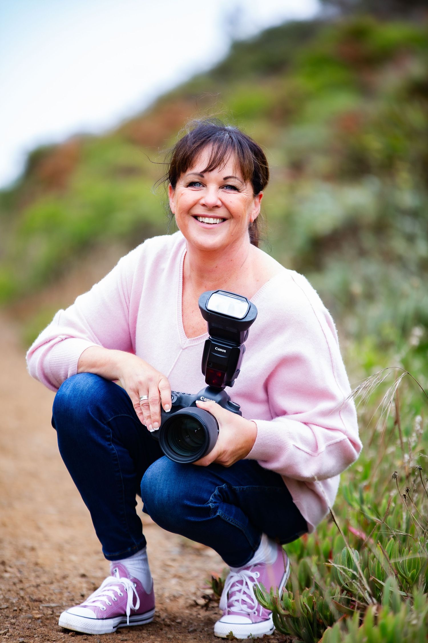 Professional photographer crouches on hiking trail wearing pink sweater and jeans while holding camera with flash attachment.